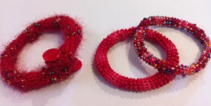 Three red bangles. Two crocheted with variegated cotton embroidery thread and a) bead mix; and b) red beads. The one on the left is crocheted with mohair yarn and cotton yarn. the beads were threaded onto the cotton yarn. Wire strengthens the structure and buttons finish it.